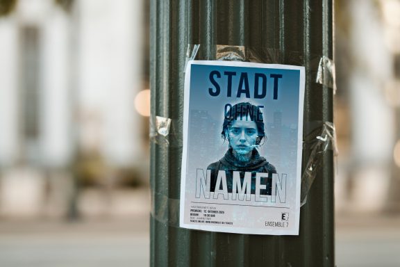 Flyer an einem Pfosten mit dem Text "STADT OHNE NAMEN" und einem Porträt einer Person.